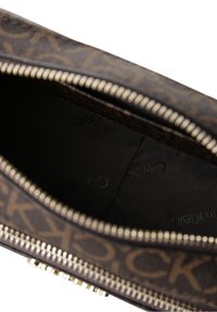 Brązowa torebka w wzory z elementami w odcieniu złota i zamkiem błyskawicznym. Wnętrze wyściełane jednolitym czarnym materiałem z delikatnym logo.