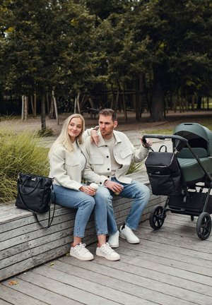 Kočárek v tmavě zelené barvě s černými textilními detaily. Dvě osoby sedí na dřevěné lavici, oblečené v světle zbarvených bundách a džínách, s černou taškou.