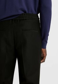 Sorte bukser med en glat overflade, der har en strækbar talje og baglommer. Matchet med en mørk lilla sweater.