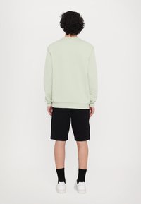 Felpa verde chiaro a collo rotondo, con una vestibilità rilassata, abbinata a pantaloni corti neri fino al ginocchio. Sneakers bianche e calzini neri completano l'outfit.