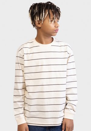 Αγόρι με κοντά dreadlocks που φοράει λευκό μπλουζάκι μακρυμάνικο με λεπτές μαύρες οριζόντιες ρίγες, κοιτώντας προς τα δεξιά του.