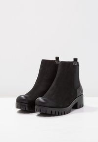 Par de botas negras hasta el tobillo con suelas gruesas, paneles laterales elásticos y tiradores en una superficie blanca con un fondo blanco liso.