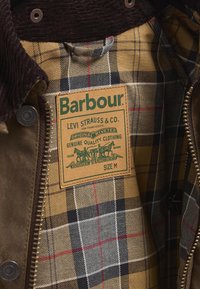 Brown jacket interior with plaid lining, featuring tan, black, and red hues. Includes a label with "Barbour" and "Levi Strauss & Co." details.