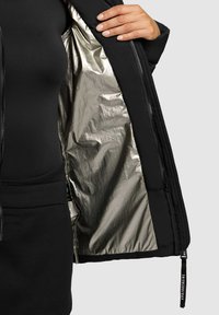 Jachetă exterioară neagră cu căptușeală interioară lucioasă argintie, având o textură netedă și închidere cu fermoar, cu un accent pe prinderea fermoarului.