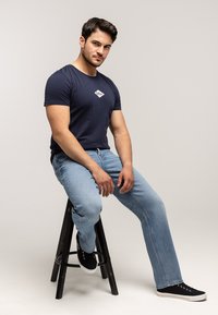 Granatowy bawełniany t-shirt z białym diamentowym logo, zestawiony z jasnoniebieskimi dżinsami i czarnymi trampkami na czarnym drewnianym stołku.