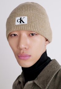 Hombre que lleva un gorro de punto beige con el logo "cK", un suéter negro de cuello alto y una chaqueta de pana verde oliva frente a un fondo plano.