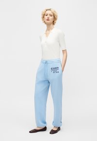 Personne portant un haut blanc à manches courtes, un pantalon de survêtement bleu clair avec des inscriptions, et des ballerines marron foncé, debout devant un fond uni.