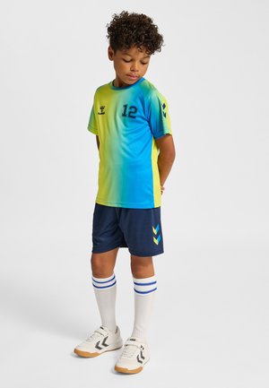 Jongen met krullend haar, gekleed in een blauw-gele sportshirt met nummer 12, donkerblauwe korte broek, witte kniekousen en witte sneakers, staand met de handen achter de rug.