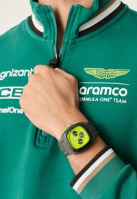 Digitālais pulkstenis ar kvadrātveida pelēku korpusu, spilgti zaļu ciparnīcu un melnu hronogrāfa subciparnīcu. Valkāts uz rokas ar zaļu kapuci un svītrainiem piedakšiem.