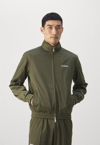 Ανδρας με ελιά πράσινο φερμουάρ ζακέτα και ταιριαστά παντελόνια, στέκεται με τα χέρια στις τσέπες, κοιτώντας μπροστά σε απλό ανοιχτό φόντο.