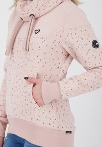 Světle růžová mikina s kapucí vyrobená z měkkého materiálu, s vícobarvnými tečkami, přední kapsou, žebrovanými manžetami a logem na rukávu.
