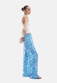 Niebieskie spodnie w szerokie nogawki z kwiatowym nadrukiem, wykonane z lekkiego materiału, z luźnym krojem i elastycznym pasem. Modelka nosi białą bluzkę.
