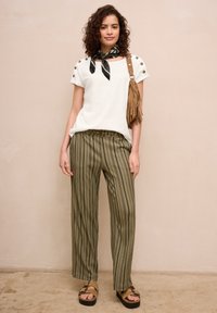 Młoda kobieta stoi, ubrana w biały top z krótkim rękawem, zielone spodnie w paski, czarną chustę na szyję, brązową torebkę na ramię z frędzlami oraz beżowe sandały na koturnie.