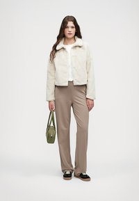 Kunstig pels kremfarget avkortet jakke med krage, sammen med beige bukser. Modellen holder en liten grønn veske, iført svarte sko med brune detaljer.