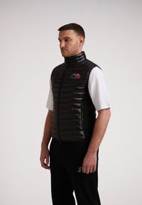 Gilet imbottito nero con motivo a righe orizzontali, colletto alto e zip frontale. Presenta un logo colorato sul petto. Indossato sopra una camicia bianca.