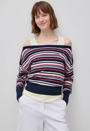 Młoda kobieta o krótkich brązowych włosach, ubrana w granatowy, biały i czerwony sweter odsłaniający ramiona, założony na żółty top i białe spodnie, z rękami w kieszeniach.