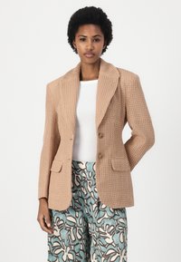 Blazer beige strutturato con revers a punte, due bottoni e tasche a patta, abbinato a pantaloni a fantasia con accenti blu e marroni.