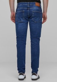 Temno modri jeans s slim fit krojem, ki imajo dva zadnja žepa in usnjeni dodatek na pasu. Gladka tekstura in minimalno šivanje.