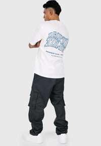 White cotton t-shirt with blue graphic text on the back, paired with dark cargo pants and white sneakers. Model is turned away from the camera.
