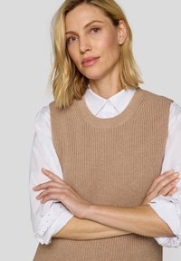 Beige gestrickte Weste mit rundem Halsausschnitt, über einem weißen Hemd mit Knöpfen getragen. Die Weste zeichnet sich durch ein strukturiertes Muster und breite Schultern aus.