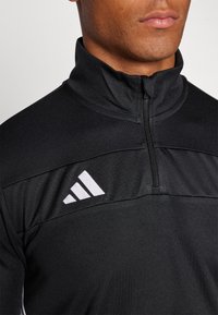Czarny sportowy polar z wysokim kołnierzem, zamek błyskawiczny i białe logo adidas. Wykonany z teksturowanej tkaniny z kontrastowymi panelami na ramionach.