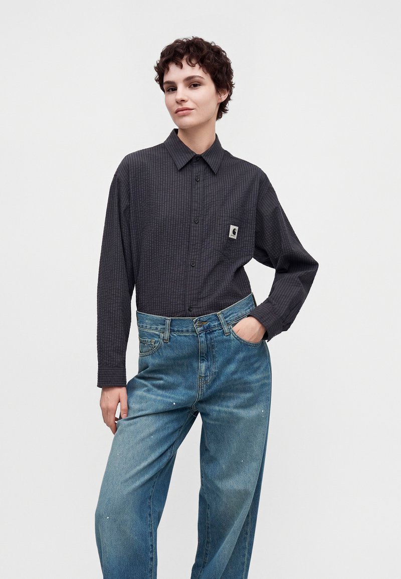 Donna che indossa una camicia a quadri scuri con bottoni e logo sulla tasca anteriore, abbinata a jeans blu larghi a vita alta, posando con una mano in tasca.