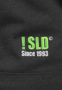 Black fabric featuring embroidered text: "! SLD" in bright green and "Since 1993" in white. Texture is soft, with a casual design.
