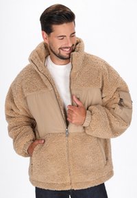 Smilende mand iført en beige plysch fleecejakke med lynlås foran og lommer, som justerer lynlåsen med den ene hånd mod en ensfarvet hvid baggrund.
