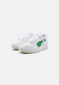 Hvite sneakers laget av lær og syntetiske materialer, med grønne detaljer og perforerte elementer, samt flate beige gummisåler.
