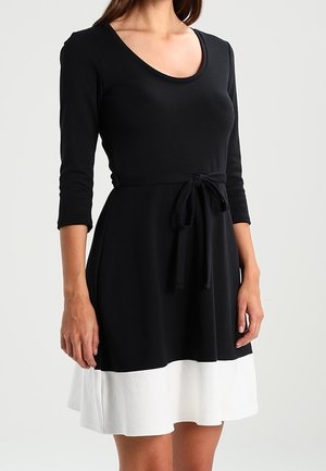 Woman wearing a black dress with three-quarter sleeves and a waist tie, featuring a white band at the hem.