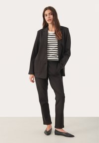 Zwarte op maat gemaakte blazer met twee voorzakken, gecombineerd met zwarte broek. Daaronder een zwart-wit gestreepte top. Zwarte platte schoenen.