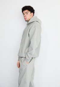 Šedá mikina s kapucňou a zodpovedajúce teplákové nohavice z mäkkej tkaniny. Obsahuje prednú kapsu a logo Nike na nohaviciach. Štíhly strih, ležérny dizajn.