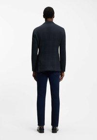 Mężczyzna stojący plecami, ubrany w ciemną marynarkę w kratę, ciemnoniebieskie dżinsy i czarne buty na jednolitym białym tle.