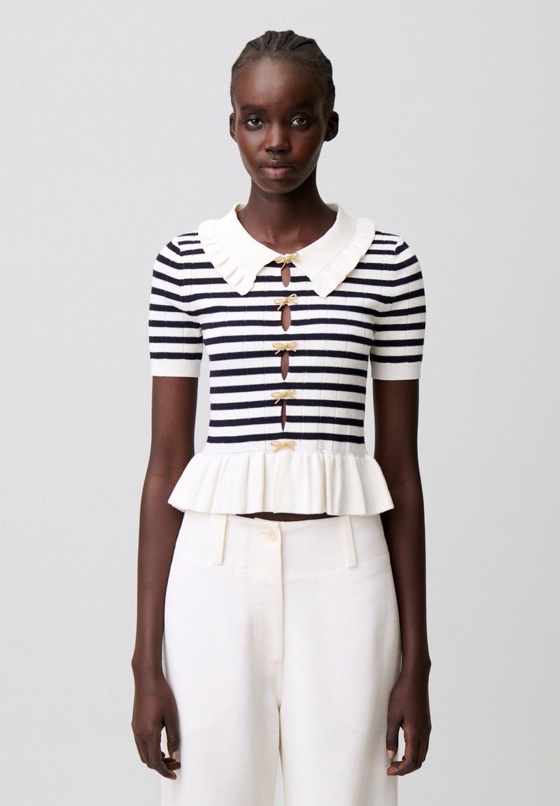 Mujer con una blusa de manga corta a rayas blancas y negras, con cuello con volantes y dobladillo con vuelo, combinada con pantalones blancos de cintura alta.