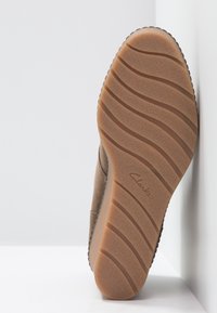 Semelle d'une chaussure beige avec des motifs ondulés et le logo "Clarks", positionnée verticalement contre un fond blanc en coin.