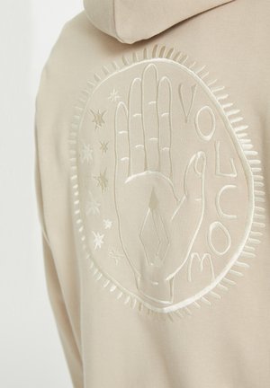 Beige hoodie featuring an embroidered hand design surrounded by stars and decorative stitching along the edges of the graphic.