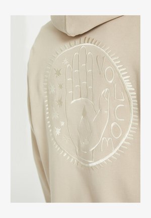 Beige hoodie featuring an embroidered hand design surrounded by stars and decorative stitching along the edges of the graphic.