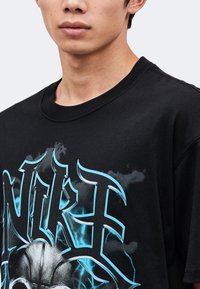 Νέος που φοράει μαύρο T-shirt με μπλε στυλιζαρισμένο κείμενο και γραφικό μπάσκετ στο μπροστινό μέρος.