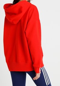 Sudadera roja de gran tamaño con capucha y textura suave. La parte trasera muestra un bolsillo canguro y puños acanalados, combinada con pantalones deportivos azul marino.