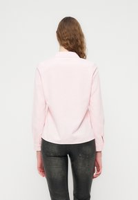 Ανοιχτό ροζ πουκάμισο με κουμπιά, μακριά μανίκια και γιακά. Το ύφασμα φαίνεται μαλακό. Φοριέται με μαύρο, εφαρμοστό παντελόνι με υφή.