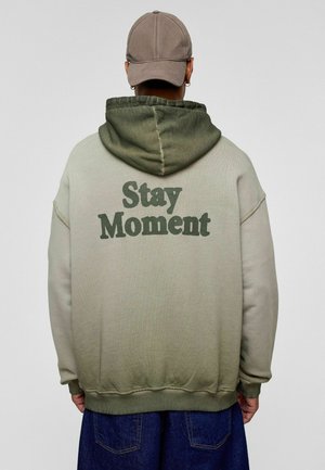 Hoody in lichtgroene gradient, met de tekst "Stay Moment" in donkergroene letters op de achterkant. Zacht materiaal, losse pasvorm en ribgebreide boorden.