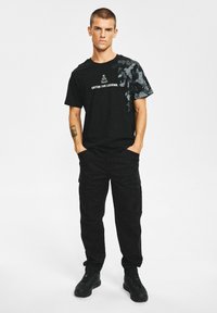 Czarny t-shirt z grafiką i napisem „ENTER THE LEGEND” z przodu, zestawiony z czarnymi spodniami typu cargo i czarnymi sneakersami.