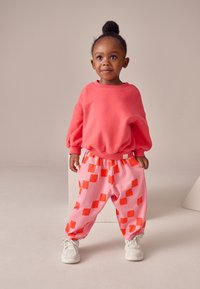 Jasno koralowa bluza z bufkami na rękawach, zestawiona z różowymi spodniami w pomarańczowe kwadraty. Dopełnieniem stroju są białe trampki.