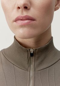 Beige svetr na zip s vysokým vroubkovaným límcem, který má kovový zip. Tkanina má texturovaný povrch a jemné vertikální pruhy.