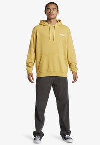 Yellow pullover hoodie with a front pocket and drawstrings, paired with dark corduroy pants and black sneakers with white stripes.