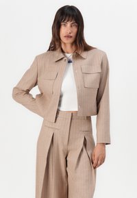 Giacca cropped a strisce marroni con due tasche sul petto, chiusura frontale a scatto e maniche lunghe, abbinata a pantaloni plissettati coordinati.
