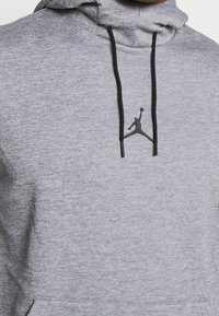 Šedá mikina s kapucí, vyrobená z texturované látky, s černým logem Jumpman na výstřihu a černými šnúrkami.