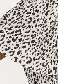 Biały materiał plisowany w czarne, nieregularne plamy w leopardzie, ukazujący część spódnicy lub sukienki na białym tle.