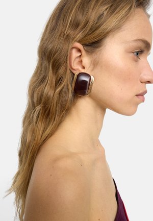 Profil kobiety z długimi, falującymi włosami, noszącej duży, prostokątny, ciemnobrązowo-złoty efektowny kolczyk na lewym uchu.