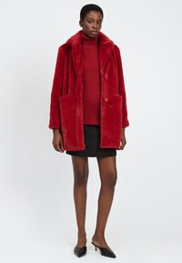 Cappotto in pelliccia sintetica rossa con ampio colletto, due tasche frontali e chiusura a doppio bottone. Indossato sopra un dolcevita coordinato e una gonna nera.
